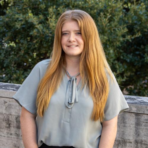 Allison has dark blonde hair. She wears a light blue flowy blouse and stands in front of tree foliage. 