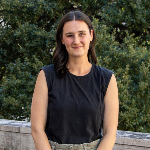 Hannah has dark brown hair. She wears a black, sleeveless shirt and stands in front of green foliage.