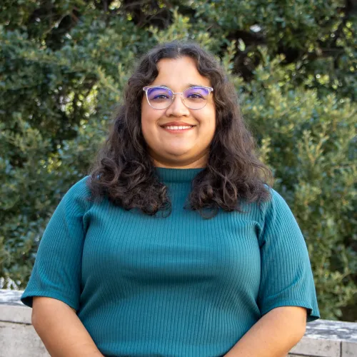 Cinthia has short, curly brown hair. She wears glasses and a teal flowy blouse.