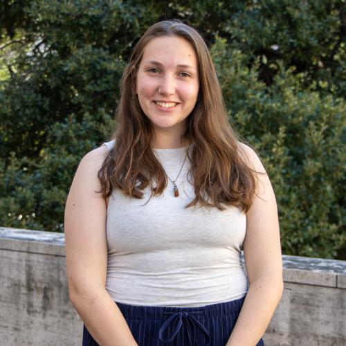 Catherine Hoffman has long brown hair. She wears an off-white sleeveless blouse and black pants and stands in front of a parapet with tree foliage in the background.