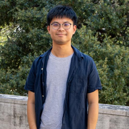 Pei-Chiang has short black hair with dark, wire glasses. He wears a grey shirt under a short sleeve navy button up shirt.