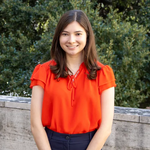 Ivana has shoulder length brown hair. She wears a red/orange blouse.