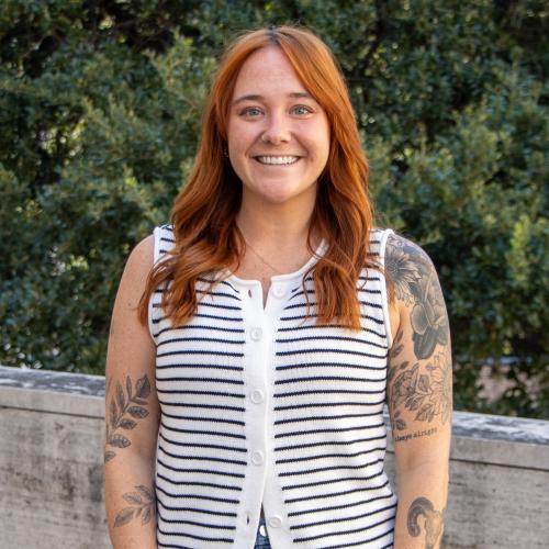 Madi has shoulder-length red hair. She wears a striped button-up blouse and has tattoos on her exposed arms. 