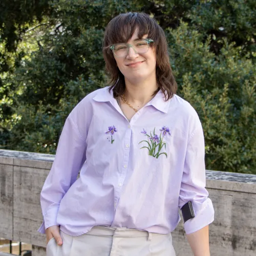 Leo has dark brown, shoulder-length hair. They wear a light purple shirt with embroidered flowers and light khakis.