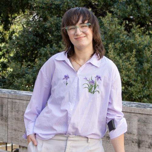 Leo has dark brown, shoulder-length hair. They wear a light purple shirt with embroidered flowers and light khakis.