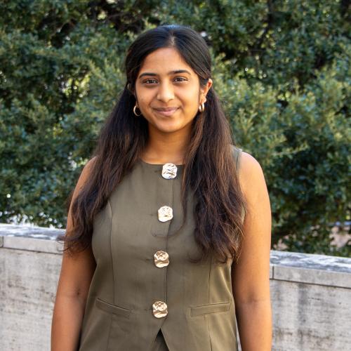Risha has long black hair. She wears an olive colored sleeveless outfit.