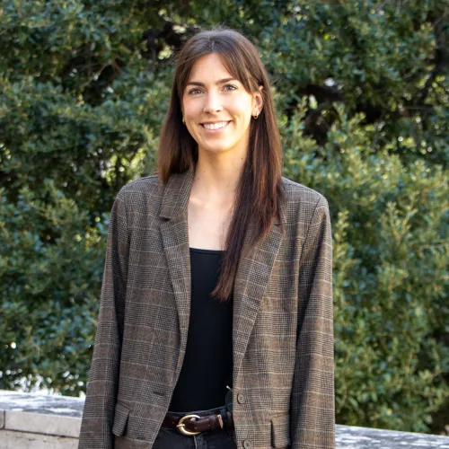 Elizabeth has straight, long brown hair. She wears a brown blazer over a black blouse.