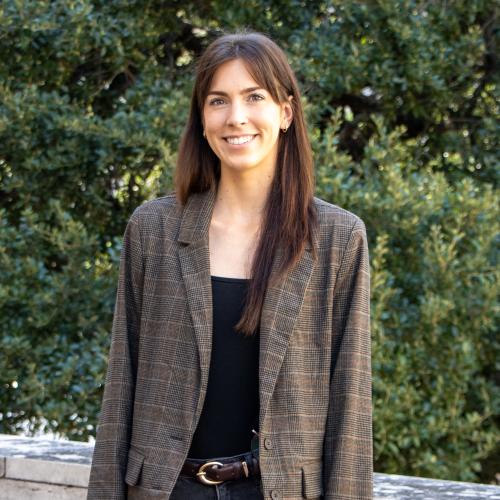 Elizabeth has straight, long brown hair. She wears a brown blazer over a black blouse.