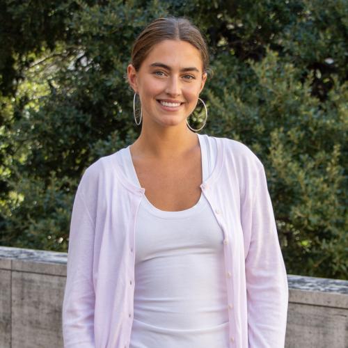 Izzy has brown hair tied back with hoop earrings. She wears a light pink cardigan and a white undershirt.