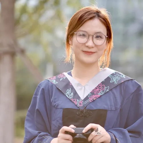Ruxin has light auburn hair. She wears round glasses and a dark purple graduation gown with a patterned collar.