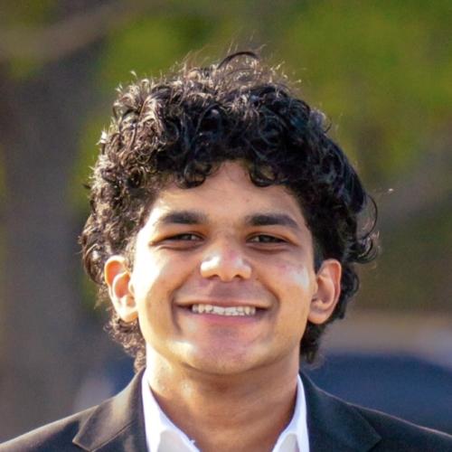headshot of Chris Kurian. Dark brown curly hair, suit on, broad smile with a wooded backdrop