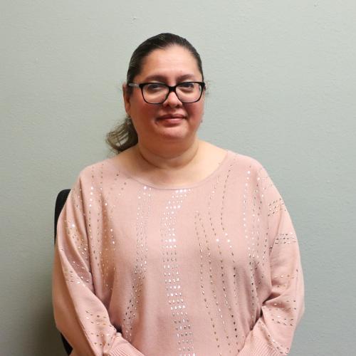 Veronica Alvarez, a woman with dark hair pulled back, wearing glasses and a pink sequined sweater, smiling gently against a plain gray-green wall.