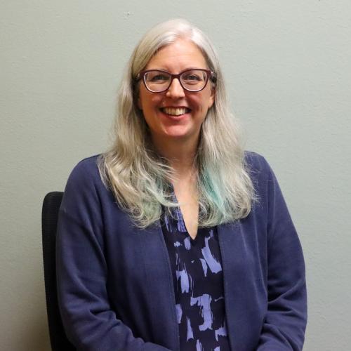 Deborah Tomai, a woman with long light gray hair with teal-tinted ends, wearing glasses and a navy cardigan over a patterned top, smiling against a plain gray-green wall.
