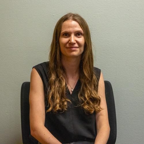 Mikayla Welch has long wavy light brown hair smiling gently, wearing a black sleeveless top with a pendant necklace, seated against a plain gray-green wall.