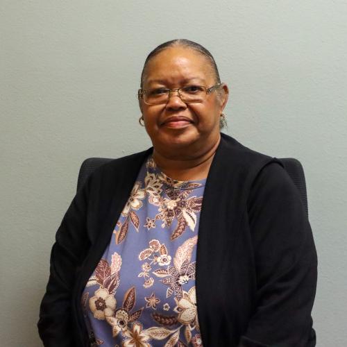 Woman with gray hair pulled back, wearing glasses and gold hoop earrings, dressed in a blue floral top with a black cardigan, seated and smiling gently against a plain light gray wall.