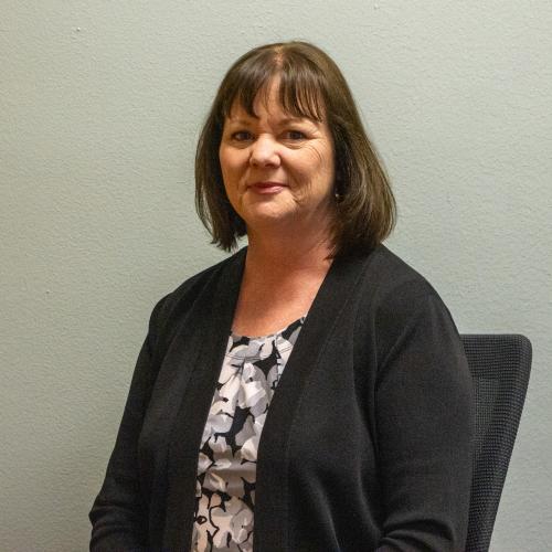 Kristen E. Davis, a woman with shoulder-length brown hair and bangs, wearing a black cardigan over a black and white floral top, smiling gently against a plain gray-green wall.