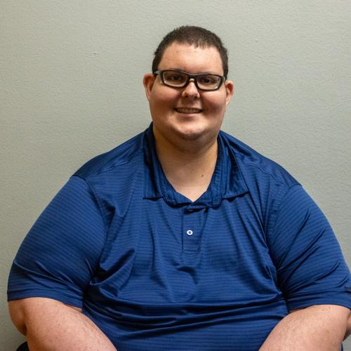 Man with short brown hair and glasses smiling in a navy blue polo shirt, seated in front of a plain light gray wall.