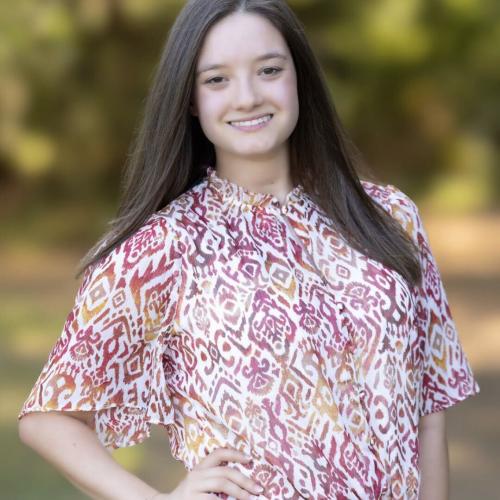 Young woman with long brown hair smiling at the camera, wearing a red, white, and gold patterned blouse tied at the waist and blue jeans, standing outdoors with a blurred green and gold foliage background, hand resting on her hip.