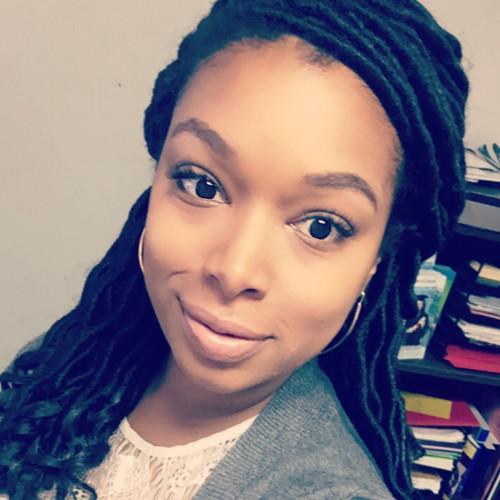 Headshot of Kristen Davis, a woman with long dark locs, smiling warmly at the camera. She's wearing a gray cardigan over a white lace top, with a bookshelf visible in the background.
