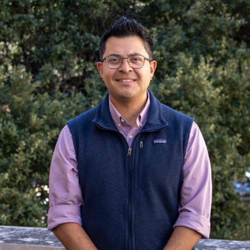 Headshot of Ricardo Bentin with short dark hair and glasses, smiling at the camera. He is wearing a navy vest over a light purple button-down shirt, standing outdoors in front of green foliage.