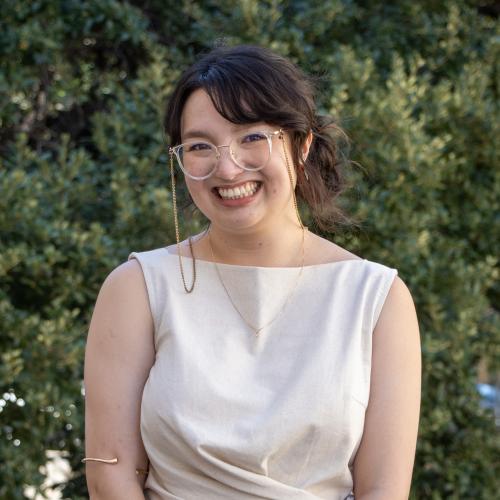 Headshot of Ashley Crooks, smiling outdoors in front of green foliage. She has dark hair pulled back, wears glasses on a gold chain, and is dressed in a cream sleeveless top.