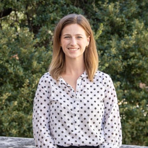 Headshot of Megan Klinginsmith, smiling outdoors in front of green foliage. She has light brown hair and is wearing a white polka-dot button-down shirt.