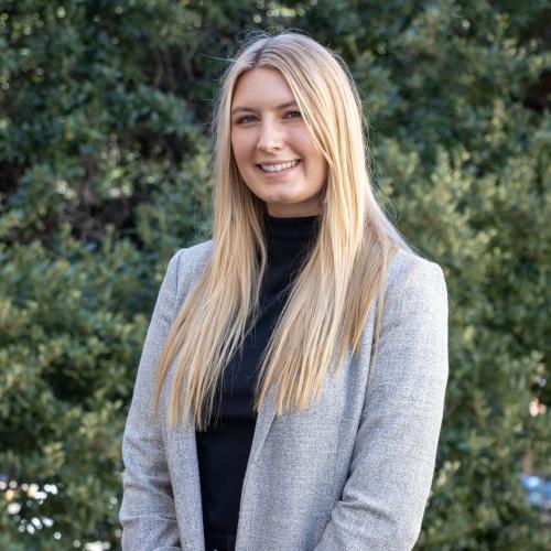 Headshot of Summer Knowles, smiling outdoors in front of green foliage. She has long blonde hair and is wearing a gray blazer over a black top.