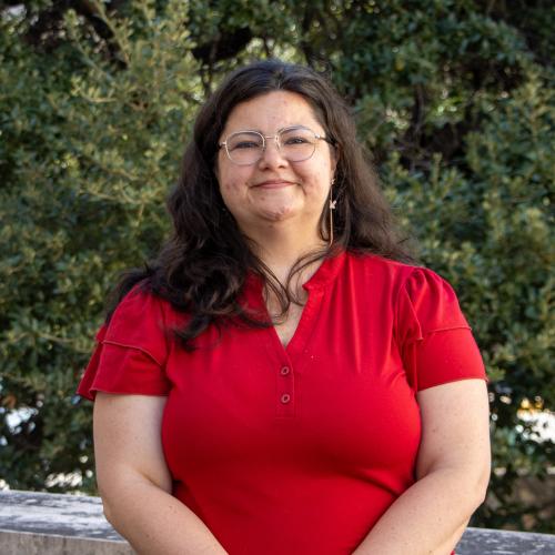 Headshot of Denise Minte de Almeida, smiling outdoors in front of green foliage. She has long dark hair, wears glasses and hoop earrings, and is dressed in a red top.