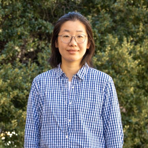 Headshot of Xian Ning, smiling outdoors in front of green foliage. She has short dark hair, wears round glasses, and is dressed in a blue and white checked button-down shirt.