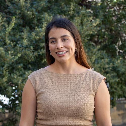 Headshot of Stephanie Soderborg, smiling outdoors in front of green foliage. She has long dark hair and is wearing a tan textured sleeveless top.