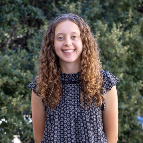 Headshot of Avery Spyers, smiling outdoors in front of green foliage. She has long curly brown hair and is wearing a navy top with a small floral print.
