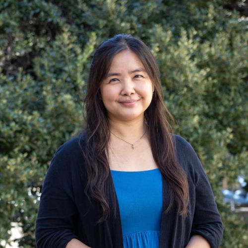 Headshot of Cindy Utama, smiling outdoors in front of green foliage. She has long dark hair and is wearing a black cardigan over a blue top.