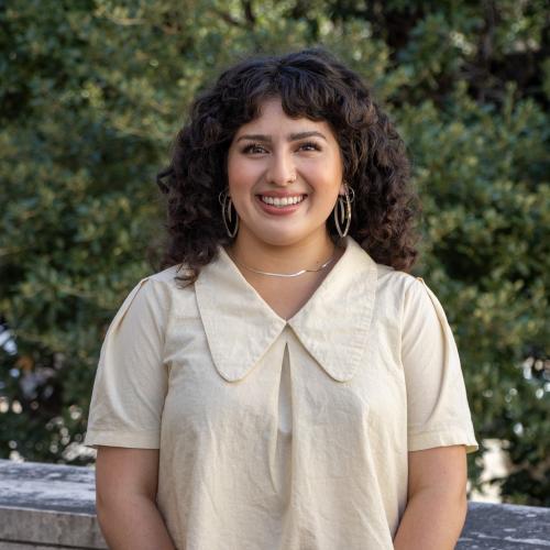 Headshot of Naiya Vasquez-Castaneda, smiling outdoors in front of green foliage. She has curly dark hair, wears hoop earrings, and is dressed in a cream blouse with a collar.
