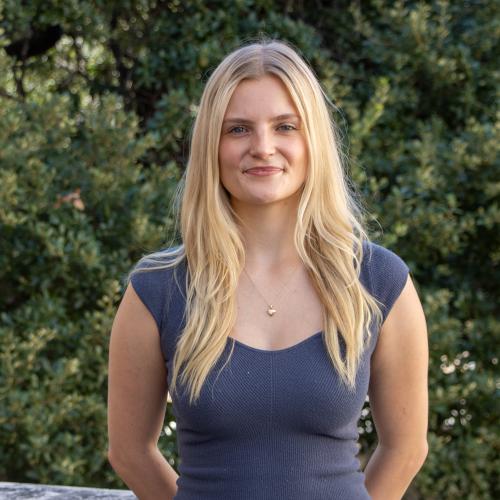 Headshot of Casey Ray Willingham, smiling outdoors in front of green foliage. She has long blonde hair and is wearing a dark gray top with a delicate necklace.