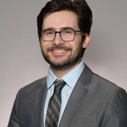 Headshot of Philip Swingon against a gray studio backdrop. He has short dark hair, a beard, and wears glasses, dressed in a gray suit with a light blue shirt and striped tie.